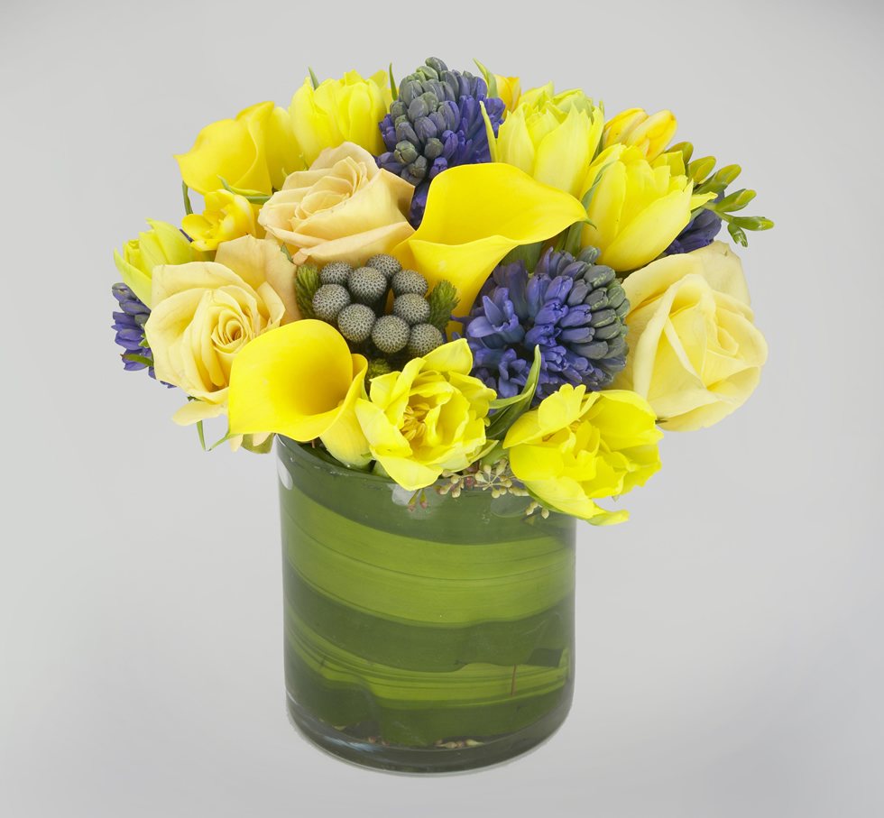 Shades of yellow and blue spring flowers with Aspidistra leaf in a cylinder vase